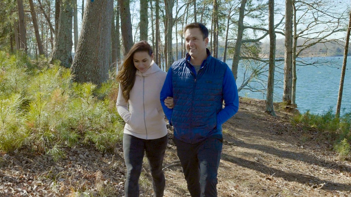 A couple walks along a wooded lakeside path on a sunny day, surrounded by tall trees and a view of the water.