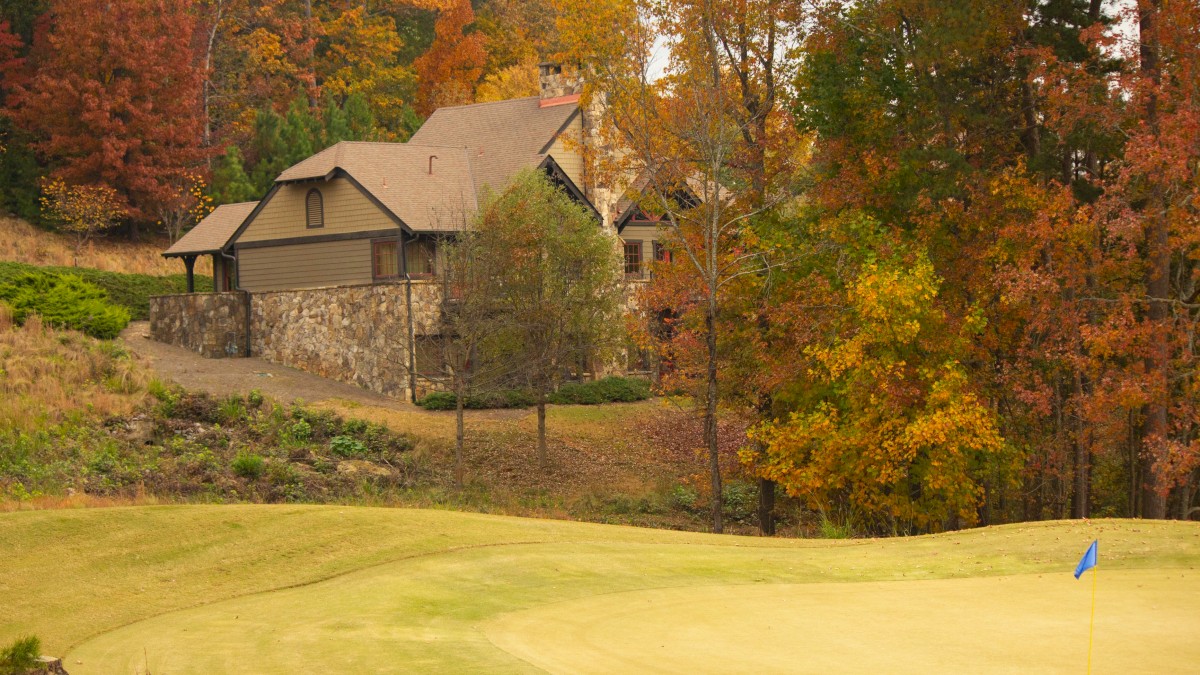 A house surrounded by autumn trees is near a golf course with a green and a blue flag, creating a serene fall landscape.