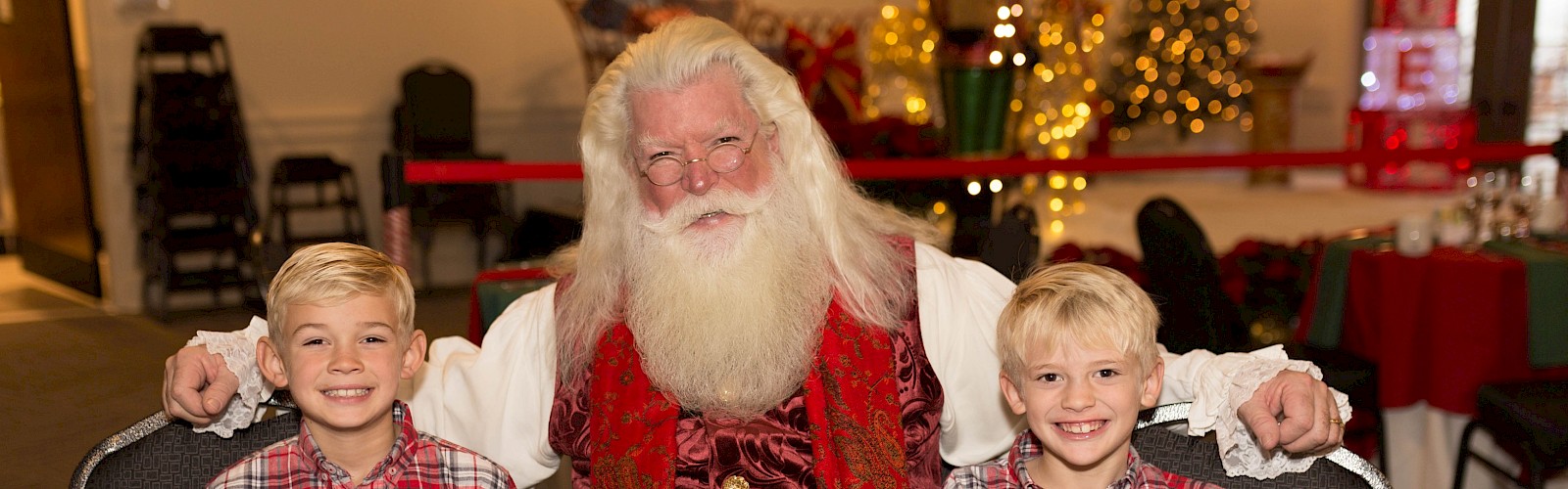 A person dressed as Santa is sitting between two children in matching plaid shirts at a festive breakfast table.