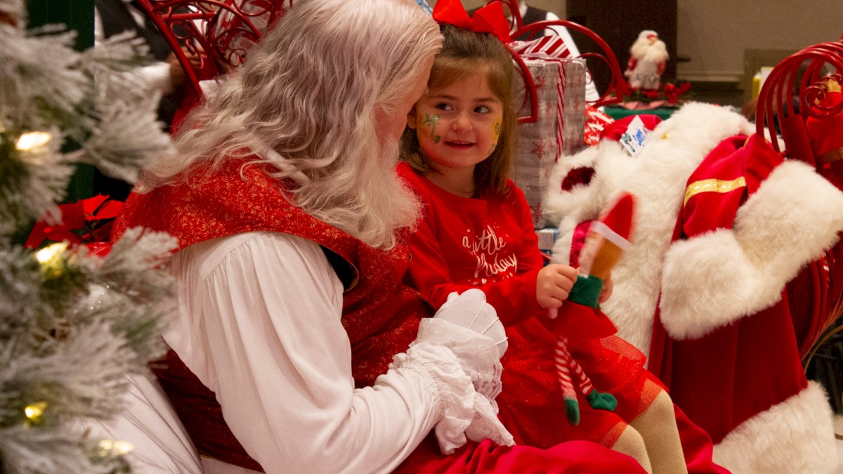 A girl in festive attire sits with Santa Claus, surrounded by Christmas decorations, enjoying a holiday moment together.