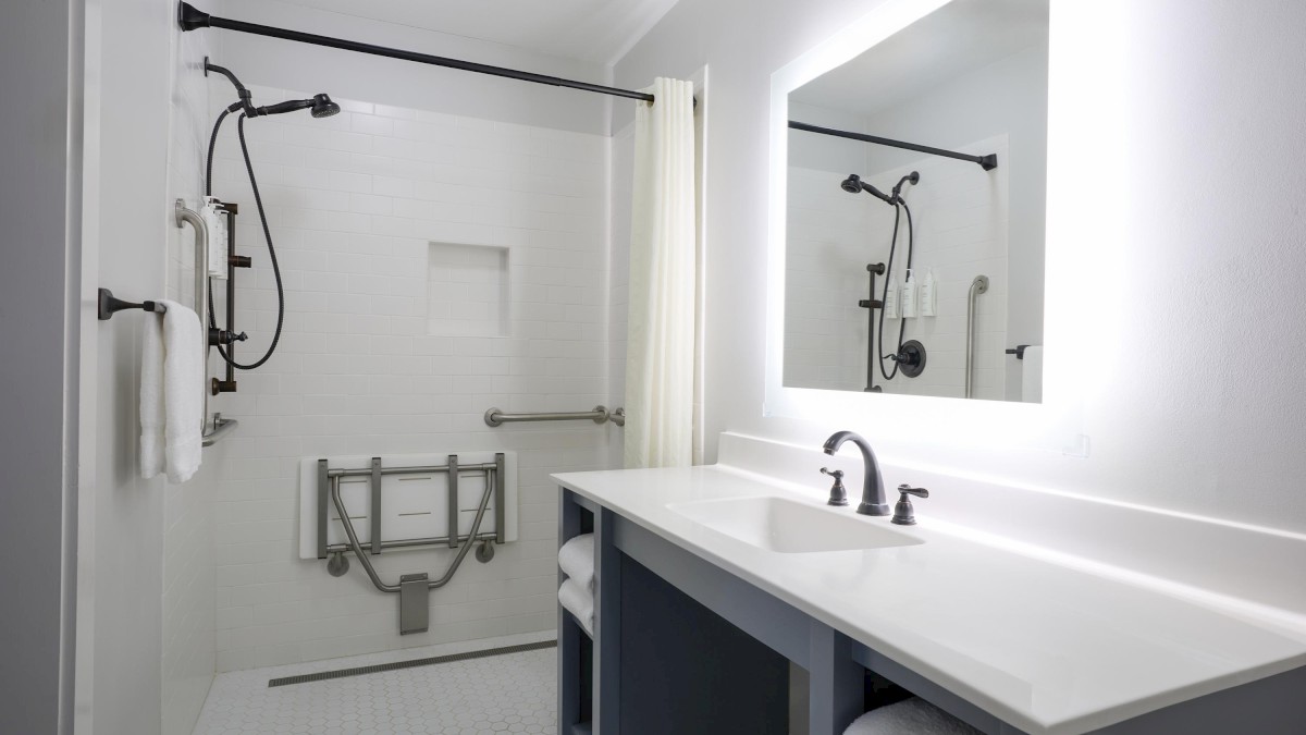 A modern bathroom with a walk-in shower, foldable shower seat, towel rack, and a wide sink with an illuminated mirror.