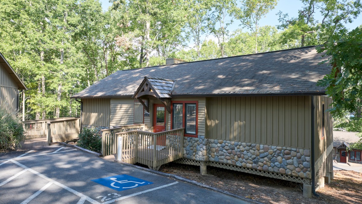 A house with a stone accent and wooden siding surrounded by trees, featuring a ramp and a handicapped parking spot in front.