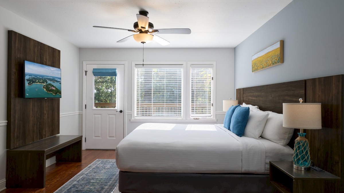 A modern bedroom with a bed, blue pillows, two lamps, a ceiling fan, wall art, and a TV. There's a window and door with blinds.