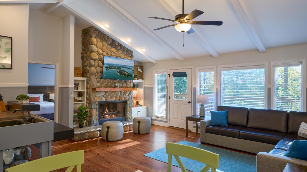 A cozy living room features a stone fireplace, ceiling fan, and large windows. The furniture is modern with a touch of rustic accents.