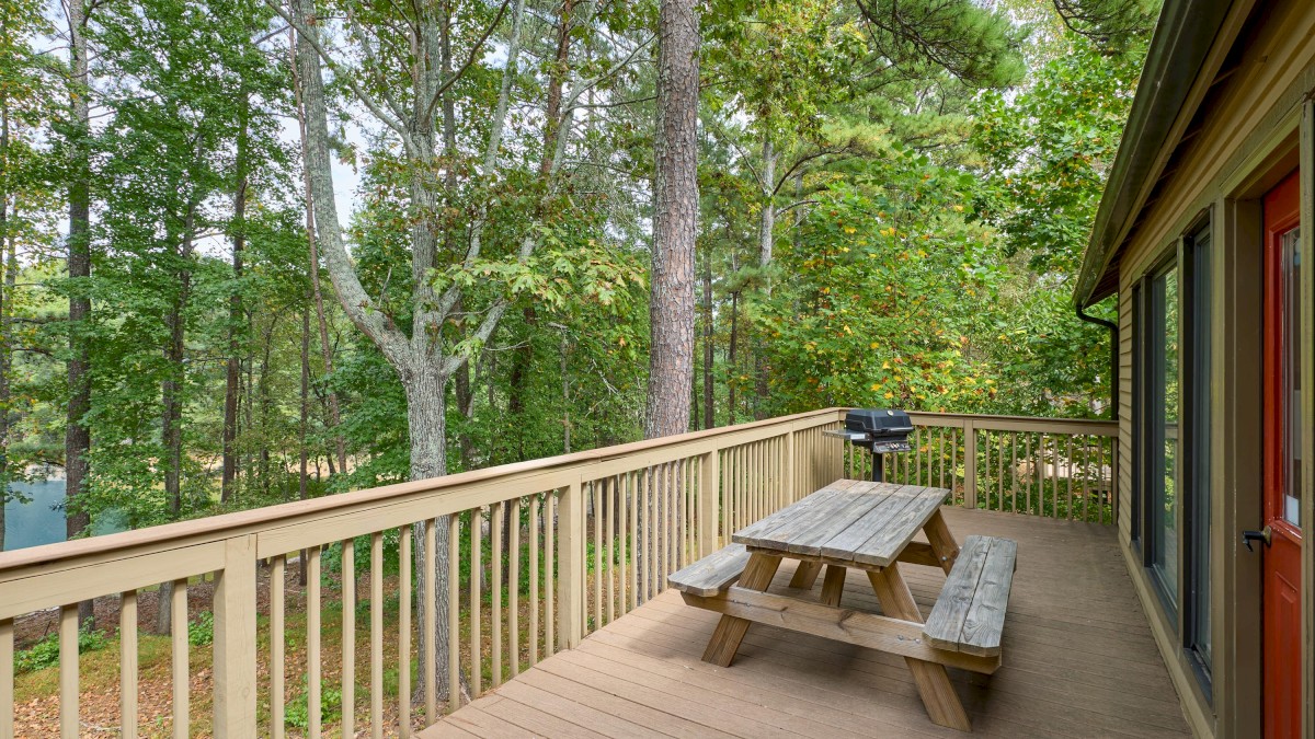A wooden deck with a picnic table overlooks a forested area, featuring a grill and a view of lush green trees.