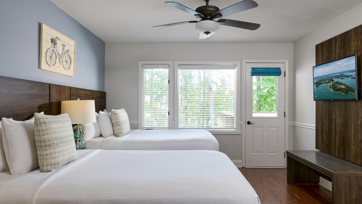 A tidy hotel room with two beds, bicycle artwork, wall TV, door, and windows with blinds; has a ceiling fan and wood flooring.