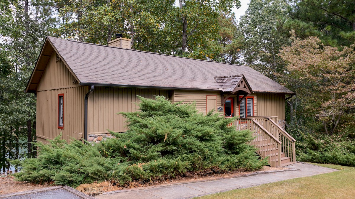 A small wooden cottage with a staircase, surrounded by trees and bushes, situated on a neatly paved and grassy area.