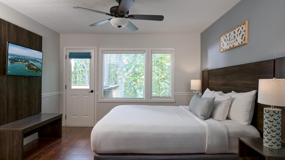 A cozy bedroom with a double bed, ceiling fan, wall-mounted TV, lamps, and a scenic view through large windows.
