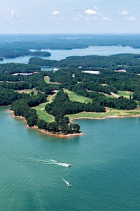 An aerial view of a large, lush lake with numerous boats and islands, surrounded by dense green forests and a bright, partly cloudy sky.
