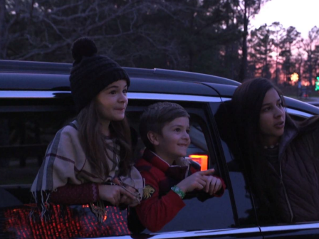 Three children are leaning out of a car window at twilight. One points toward something outside, framed by trees and a softly lit sky.