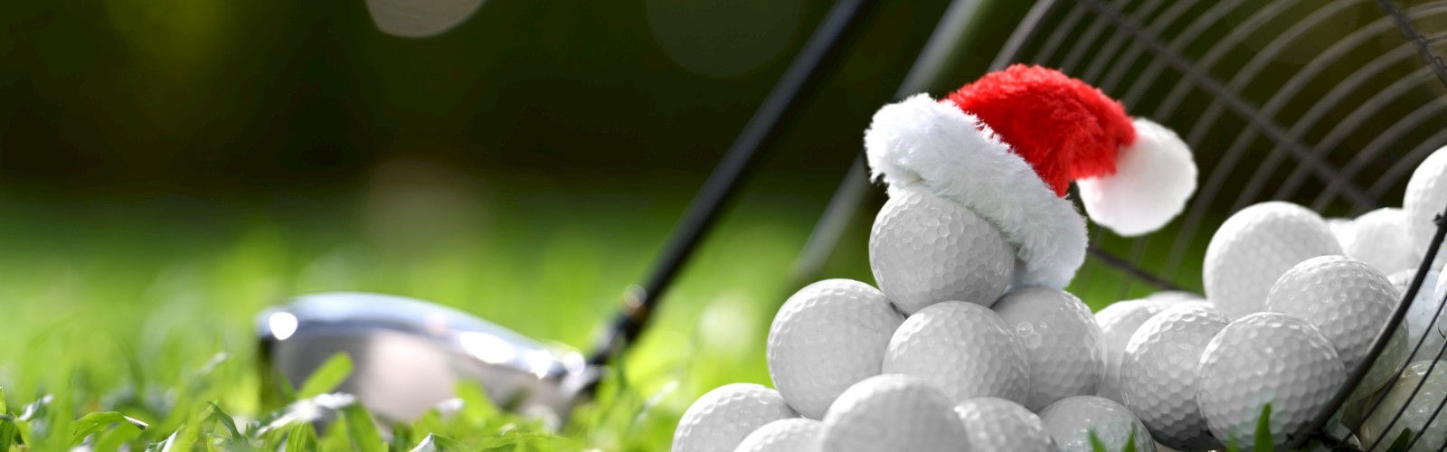 A pile of golf balls with a small Santa hat, a golf club, and a basket on grass, with a blurred green background.