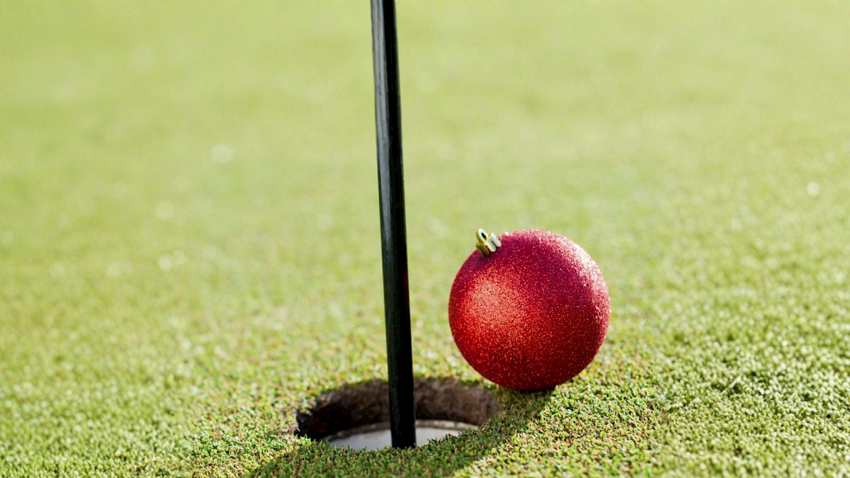 A red Christmas ornament rests beside a golf hole flag on a green course, casting a shadow.