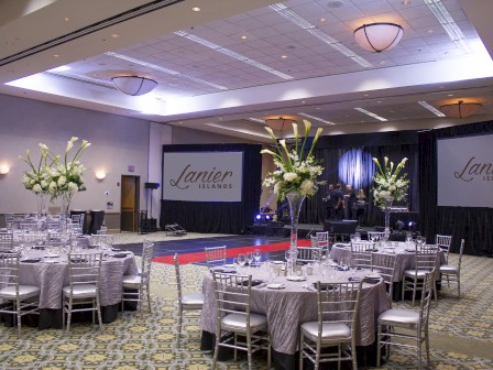 The image shows an elegant banquet hall setup with round tables, flower centerpieces, and a stage with 