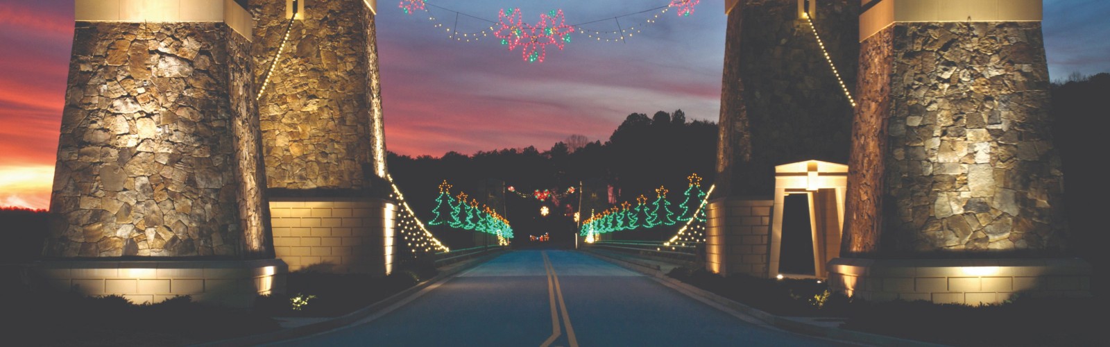 A road entrance with stone towers, adorned with festive lights and trees, set against a colorful dusk sky.