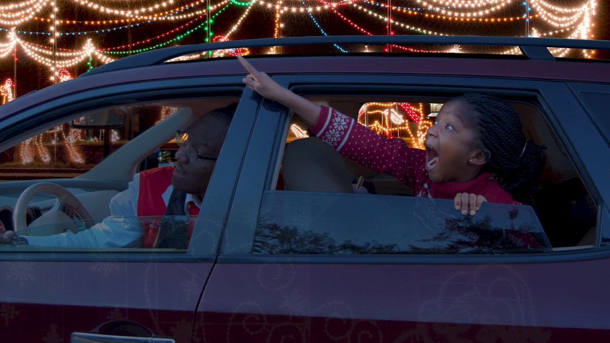 A child excitedly points out Christmas lights while leaning out of a car window at night.