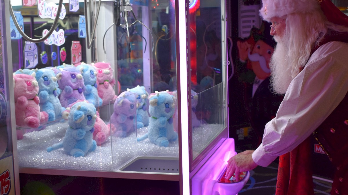A person dressed as Santa is playing a claw machine filled with stuffed toys at a venue.