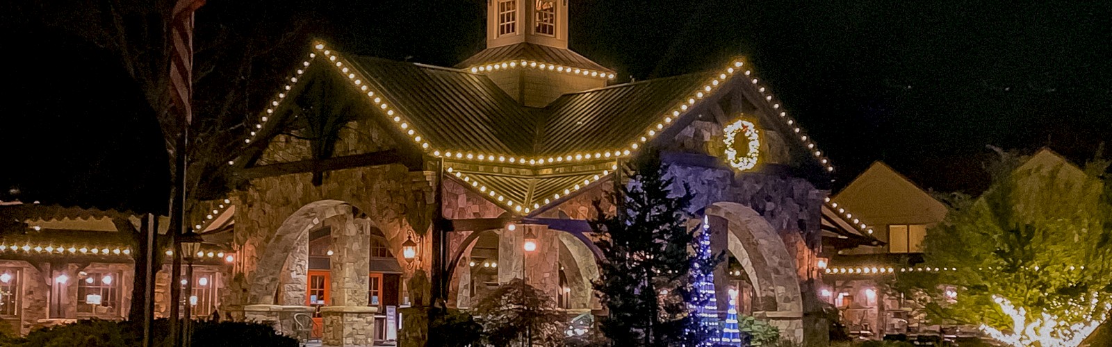 A building adorned with festive lights and a wreath at night, surrounded by trees and a decorated pathway, creates a cozy atmosphere.