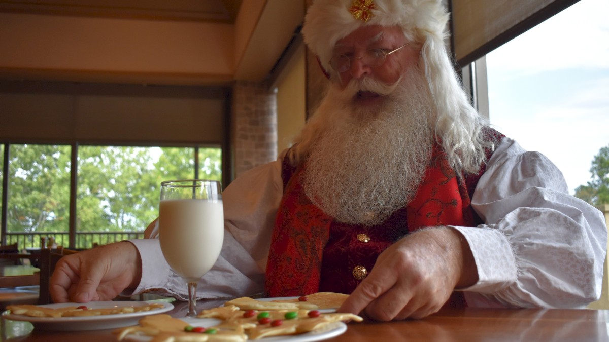 A man dressed as Santa Claus is sitting at a table with plates of cookies and a glass of milk.