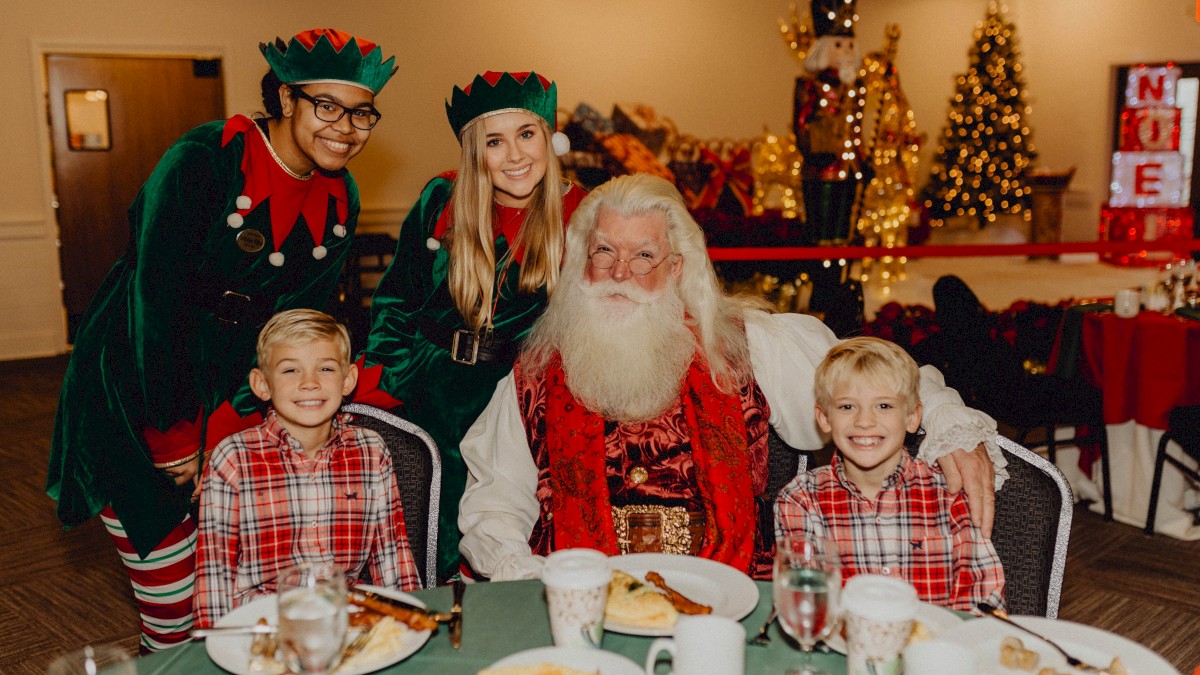 A festive scene with Santa, two elves, and two children at a holiday table, surrounded by Christmas decorations and trees.