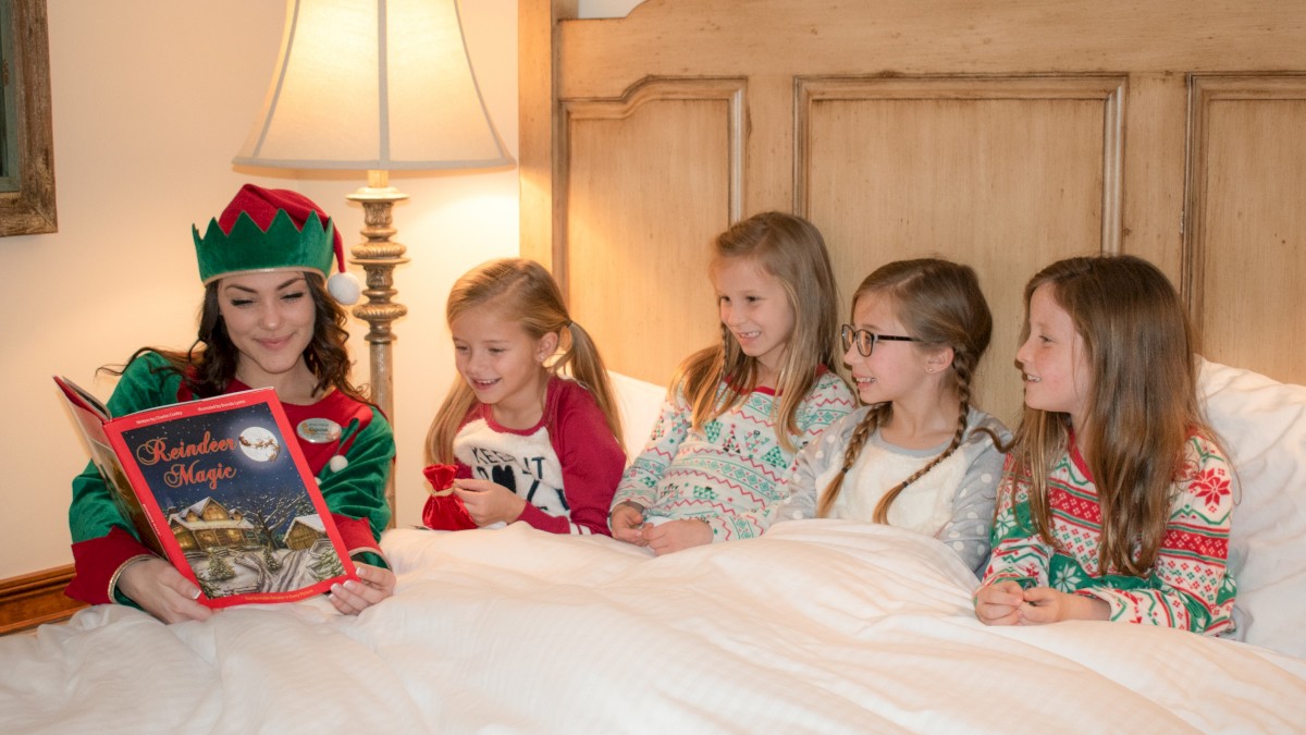 A person dressed as an elf reads a book to four children in a bed, with a lamp beside them for lighting.