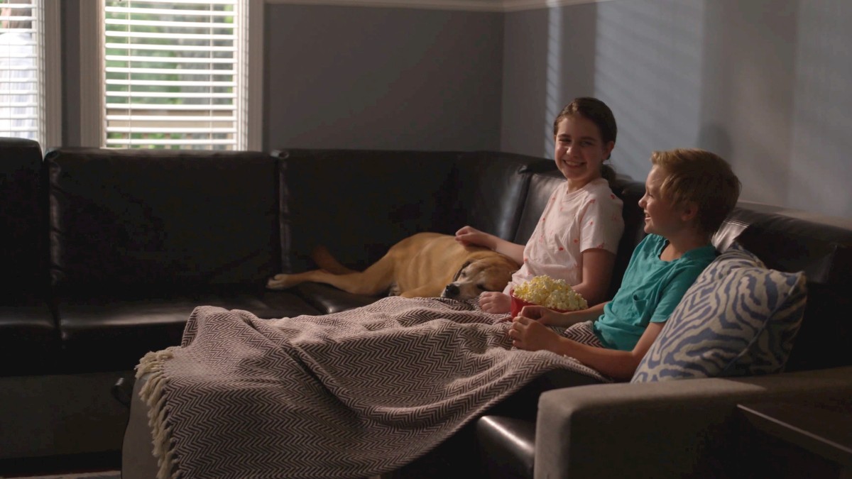 A dog and two people relaxing on a sofa, sharing a blanket and enjoying snacks.