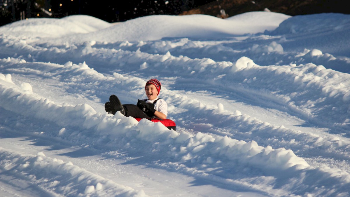 A child is sliding down a snowy hill on a sled, joyfully wearing winter clothing amidst deep snow tracks, with trees in the background.