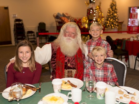 A person dressed as Santa with three smiling children at a festive table, featuring Christmas decorations in the background.