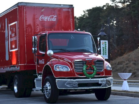 A red Coca-Cola truck is parked outdoors, decorated with a festive wreath on the grill, beside a table and heater.