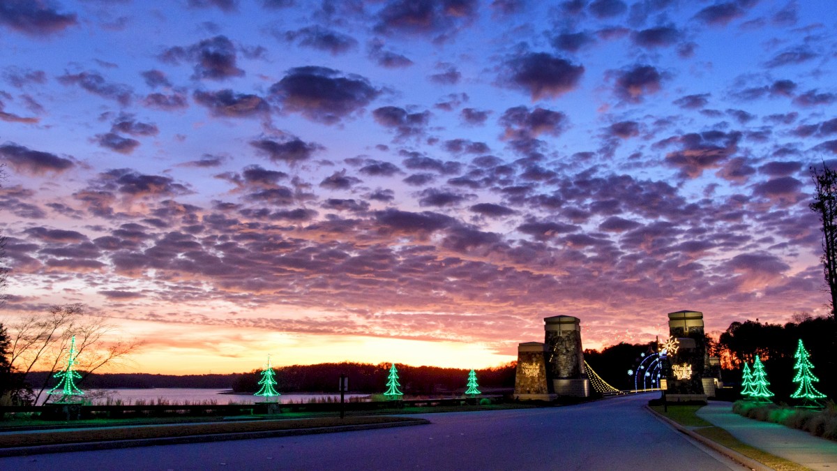 A scenic sunset over a bridge with illuminated trees lining the road beneath a dramatic sky filled with scattered clouds.