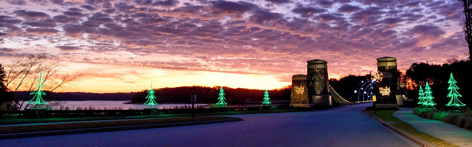 A sunset sky with clouds, a lit bridge, and glowing tree decorations along the road, creating a festive scene.
