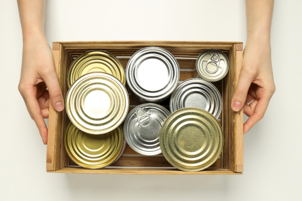 A person holds a wooden box filled with various sealed metal cans, arranged neatly inside.