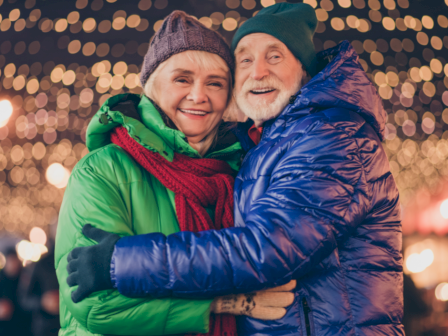 An elderly couple in winter clothing embraces, smiling under festive lights in a joyful, outdoor setting.