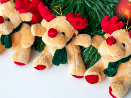 Four plush reindeer ornaments with red noses and antlers, wearing green scarves, are arranged near pine branches and red baubles.