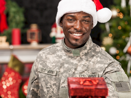 A person in military uniform with a Santa hat is smiling and holding a gift box, with a festive background.