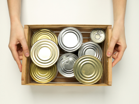 A pair of hands holding a wooden crate filled with a variety of metallic canned goods, viewed from above.