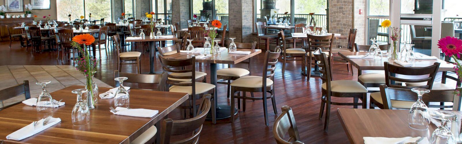 A spacious, modern restaurant with wooden tables and chairs, set with cutlery, glasses, and flowers in vases, features large windows and natural light.