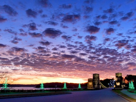 A scenic sunset with a bridge, illuminated trees, and a vibrant sky with fluffy clouds reflecting various colors.