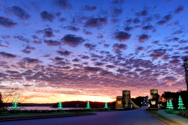 A scenic sunset with a bridge, illuminated trees, and a vibrant sky with fluffy clouds reflecting various colors.