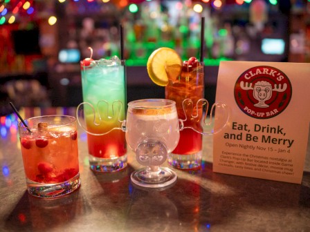 Assorted colorful cocktails on a bar counter with a Clark’s joint card reading “Eat, Drink, and Be Merry.”