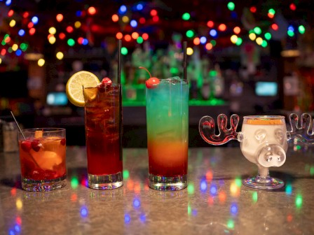 Three colorful cocktails on a bar counter with festive lights in the background, a lime wedge, and a quirky glass on the right.
