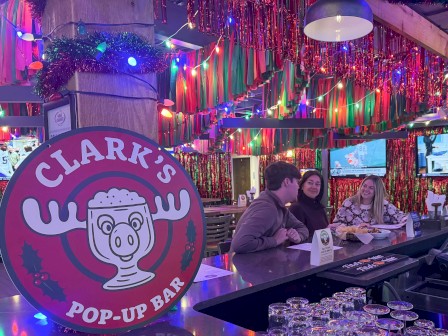 A colorful pop-up bar with festive lights, neon colors, and a Clark’s Pop-Up Bar sign featuring a cartoon moose, people chatting at the counter.