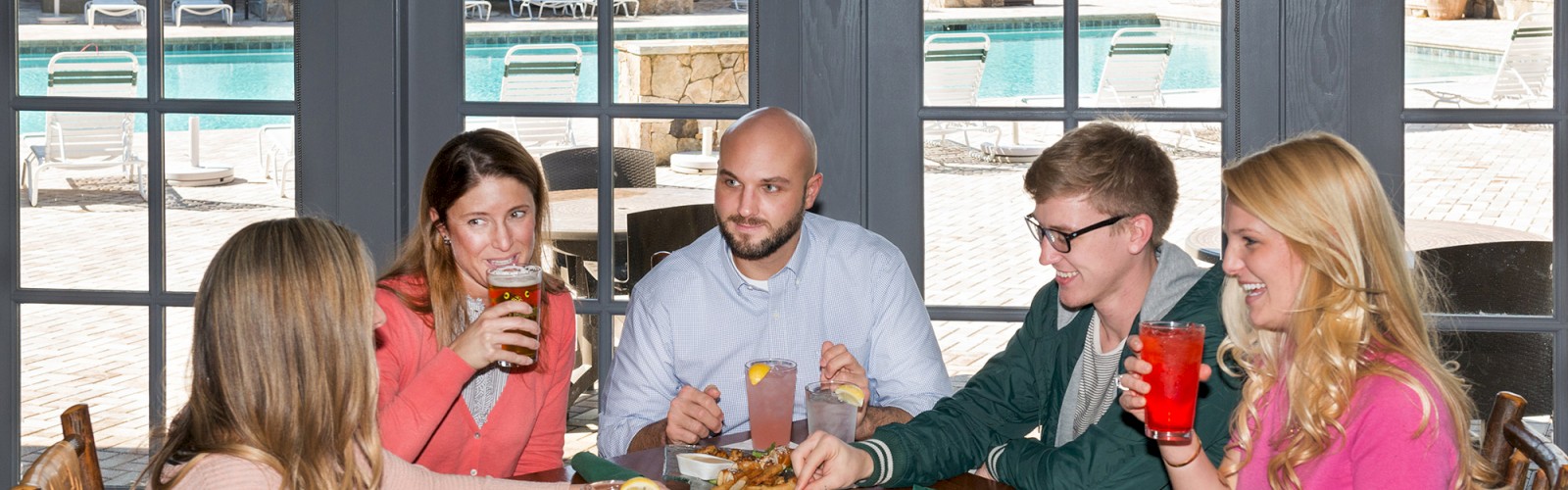 A group of five people enjoying food and drinks around a wooden table indoors, with a pool visible in the background through large windows.
