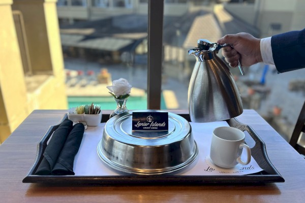 A hotel breakfast setup on a tray: metal teapot being poured, cup, napkins, sugar, and a small plant against a window with urban backdrop.