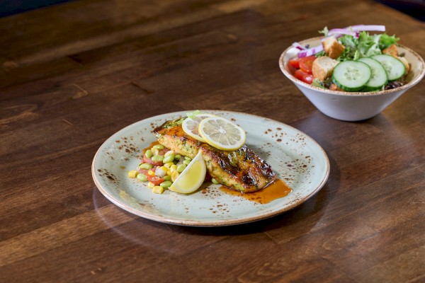 A plated meal with fish topped by a lemon slice, served with vegetables and a wedge of lemon on a rustic plate, plus a side salad in a bowl.