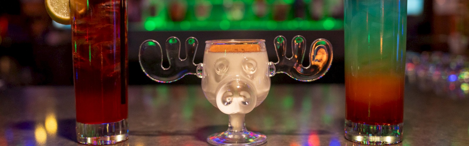 Three colorful cocktails on a bar counter with a candleholder face in the middle, festive lights glowing in the background, cheers.