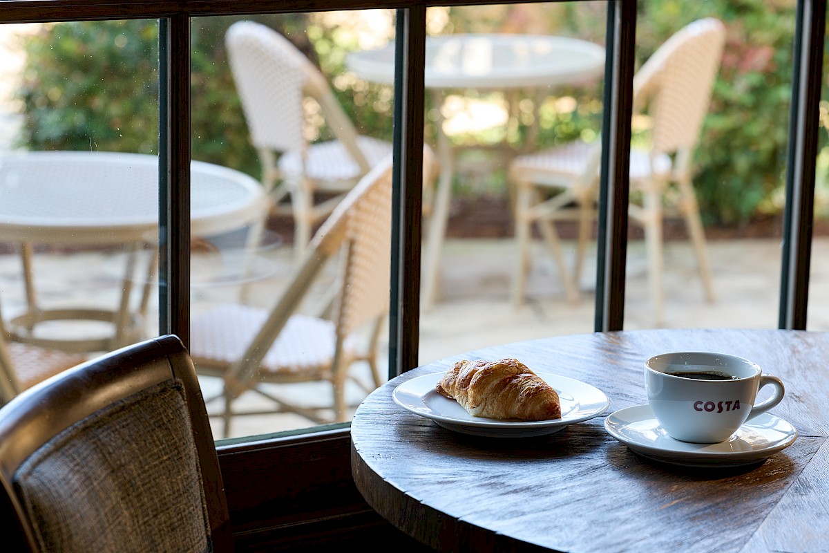 A croissant on a plate with a cup of coffee on a wooden table by a window, outdoor chairs visible outside.
