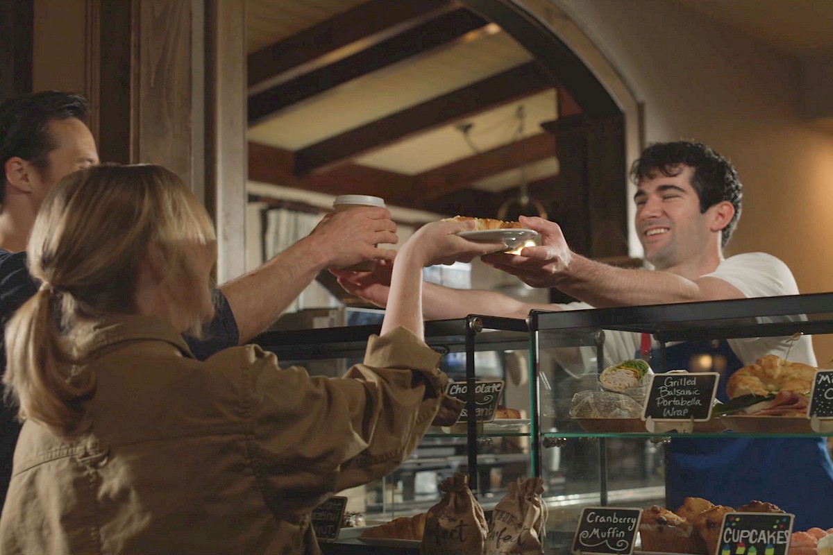 People at a bakery counter raise their glasses to toast a seller behind the glass display, a cheerful moment of celebration.