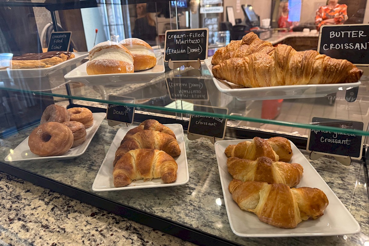 Assorted pastries in a bakery display: croissants, pain au chocolat, doughnuts, and rolls on white trays behind glass. End.