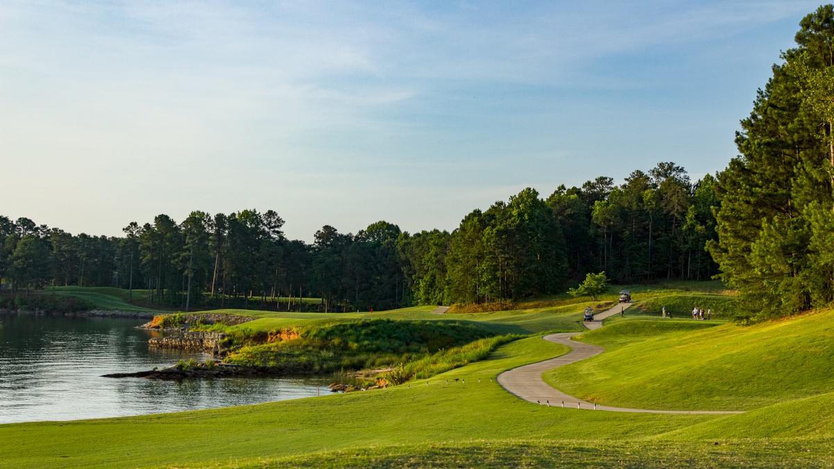 A serene lakeside golf course with a winding cart path, lush green fairways, and trees lining the water under a clear blue sky.