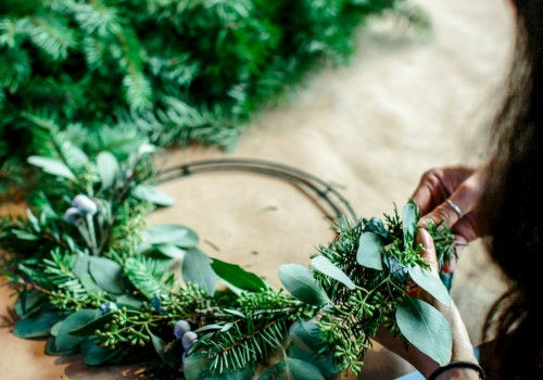 A handmade wreath being crafted with greenery and eucalyptus, by a person’s hands on a wooden table, with tools and foliage nearby.